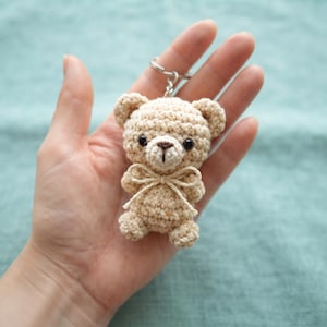 Op de afbeelding: Een kleine, beige, gehaakte teddybeer-sleutelhanger met een rond hoofd, twee kleine oortjes, donkerbruine plastic ogen, een geborduurde neus en een dunne, lichtgekleurde strik van touw om de nek, bevestigd aan een zilverkleurige metalen ring.