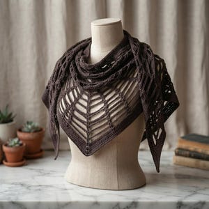 May include: A dark brown triangular crochet shawl featuring an intricate openwork web-like pattern. The garment is draped over a beige mannequin, showcasing the detailed yarn construction, pointed edges, and the textured stitch work throughout the piece.
