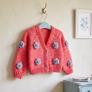 May include: A coral-colored crochet cardigan featuring a V-neckline, long sleeves, and three purple buttons. The garment is decorated with several light blue, three-dimensional flower appliqués attached to the front and sleeves.