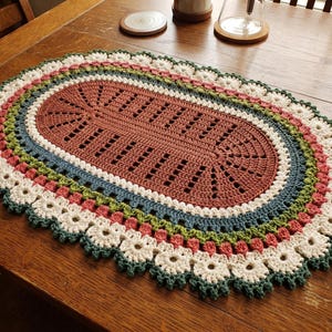 May include: An oval crochet doily featuring a terracotta center with a geometric cutout pattern, surrounded by concentric rings of blue, green, and pink yarn, finished with a scalloped white and dark green floral-inspired border edge.