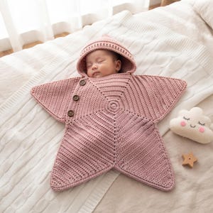 Può includere: Una coperta avvolgente per neonati all'uncinetto color rosa polvere a forma di stella, dotata di cappuccio e tre bottoni marroni sul lato. La coperta presenta un motivo a maglia testurizzato progettato per avvolgere il neonato in modo sicuro.