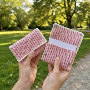 Puede incluir: Dos estuches rectangulares de ganchillo hechos de hilo rosa claro con un borde blanco. Un estuche está cerrado, mientras que el otro está abierto, revelando una tarjeta rectangular blanca en su interior. Ambos estuches tienen botones de presión de metal plateado.