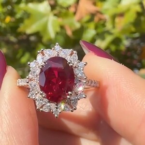 Puede incluir: Un anillo con una gran piedra preciosa de color rojo intenso y corte ovalado en el centro, rodeada por un halo de piedras blancas transparentes de corte marquesa y redondo. El aro es metálico y tiene pequeñas piedras incrustadas.