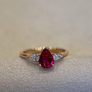 May include: A gold ring featuring a central pear-shaped deep red gemstone held in a prong setting. The ring is accented on each side by two smaller pear-shaped clear stones, creating a three-stone design with a polished gold band.