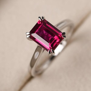 Op de afbeelding: Een zilverkleurige ring met een grote, rechthoekige, smaragdgeslepen felrode edelsteen die op zijn plaats wordt gehouden door vier opvallende pootjes. De ringband is glad en gepolijst, reflecteert het licht en benadrukt de dieprode kleur van de steen.