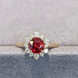 Peut inclure: Une bague en or avec une pierre précieuse centrale ronde d'un rouge profond, entourée d'un halo de douze petits diamants blancs ronds et clairs. La monture est en forme de fleur, avec la pierre rouge au centre et les diamants en pétales.