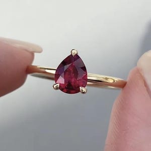 Pode incluir: Um anel de ouro com uma pedra preciosa vermelho-escura em forma de pera, fixada numa cravação de três garras. O aro é liso e polido, apresentando um design simples que destaca o corte facetado da pedra central e o tom quente do metal.