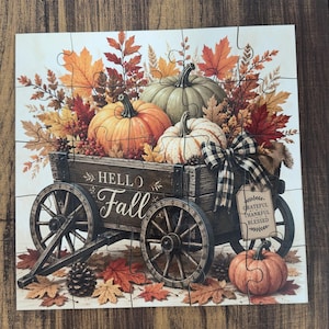 May include: A jigsaw puzzle featuring a wooden wagon filled with orange, white, and green pumpkins, autumn leaves, and pinecones. The wagon side displays the text Hello Fall, and a hanging tag reads Grateful Thankful Blessed with a plaid bow.