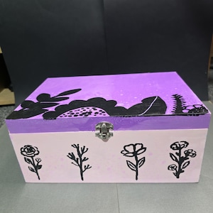 May include: A rectangular wooden box with a hinged lid and a metal latch. The lid is painted bright purple with black floral silhouettes. The front panel is light pink and features four distinct black hand-painted floral designs in a row.