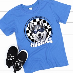 May include: A light blue t-shirt featuring a graphic of a smiling husky wearing sunglasses, set within a black and white checkered circle. The word "Huskies" is written in white below the graphic. Black sneakers with white soles are also visible.