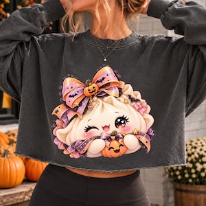 Peut inclure: Un sweat-shirt court gris anthracite orné d'un motif représentant un personnage de boulette souriant et faisant un clin d'œil avec de grands yeux. Le personnage porte un grand nœud violet et orange, des petites chauves-souris et des étoiles.