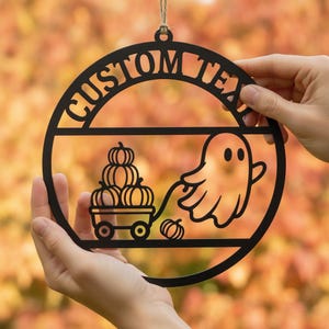 Könnte beinhalten: Ein kreisförmiges, schwarzes Hängeornament mit dem Text CUSTOM TEXT am oberen Rand. Das Design zeigt ein Gespenst, das einen kleinen Wagen mit drei gestapelten Kürbissen zieht, sowie einen weiteren einzelnen Kürbis neben dem Wagen.