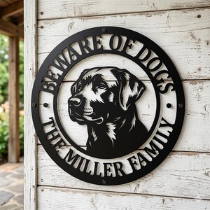 Può includere: Un'insegna circolare in metallo nero con un'illustrazione ritagliata della testa di un cane al centro. Il testo "BEWARE OF DOGS" è arcuato nella parte superiore e "THE MILLER FAMILY" in quella inferiore, entrambi in un carattere grassetto sans-serif.