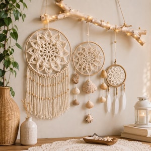 Op de afbeelding: Drie crèmekleurige gehaakte mandala's hangend aan een lichtgekleurde houten tak versierd met lichtsnoeren. De mandala's hebben ingewikkelde patronen, houten kralen, hangende schelpen en witte veren, wat een gelaagde wandhanger vormt.