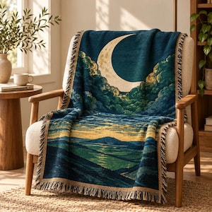 Peut inclure: Un plaid tissé représentant un croissant de lune sur un ciel bleu foncé avec des nuages verts, se transformant en un paysage de collines vallonnées et un horizon doré. La couverture présente un bord à franges et des tons bleu et vert.