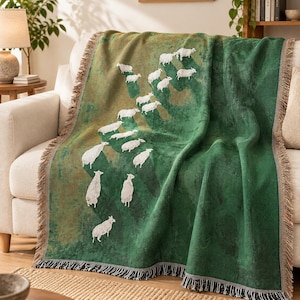 May include: A woven throw blanket featuring a green and olive gradient background with a repeating pattern of white sheep. The blanket has a textured surface and finished edges with a short, multi-colored fringe along the sides and bottom.