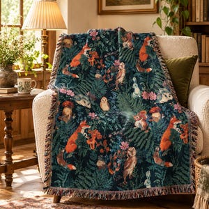 May include: A woven throw blanket featuring a dark navy background with a repeating pattern of orange foxes, brown owls, squirrels, rabbits, hedgehogs, mushrooms, and green ferns. The blanket has a decorative fringed edge on all sides.