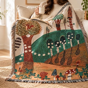 May include: A woven throw blanket featuring a colourful forest scene with a large tree, a squirrel, tall trees, and various mushrooms on a light background. The blanket has a fringed edge and displays a detailed illustration of a woodland landscape.