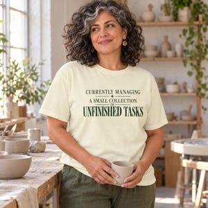 Op de afbeelding: Een crèmekleurig T-shirt met korte mouwen en donkergroene tekst die luidt: CURRENTLY MANAGING A SMALL COLLECTION OF UNFINISHED TASKS. Het T-shirt heeft een ronde hals en een relaxte pasvorm, gemaakt van een effen stof met een gladde textuur.