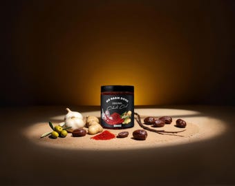 May include: A black jar labeled No Harm Done Original Chili Oil, surrounded by garlic bulbs, green olives, brown nuts, and a small pile of red chili powder. The label features illustrations of garlic, red chili peppers, and ginger root.