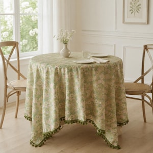 Pode incluir: Uma toalha de mesa redonda com um padrão floral em verde claro e rosa suave, com uma borda decorativa de pompons em verde escuro. O tecido cobre uma mesa circular, onde estão dispostos um vaso de cerâmica branca e dois pratos.