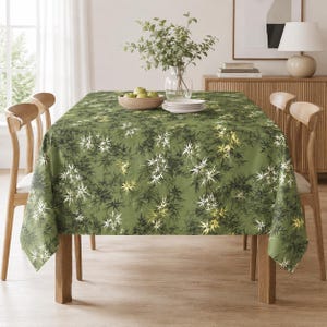 May include: A rectangular tablecloth in a muted olive green shade featuring a repeating botanical print of dark green and white maple-like leaves. The fabric drapes over a wooden table, which is set with a glass vase of greenery and plates.