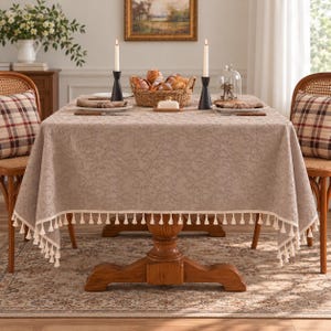 May include: A rectangular beige tablecloth with a textured floral pattern and cream-colored tassel fringe trim along the edges. The fabric drapes over a wooden table, covering the surface completely while hanging down to reveal the decorative tassels.