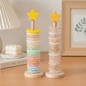 May include: Two wooden stands with round bases and vertical poles topped with yellow stars. One stand holds a stack of multicolored spiral hair ties in pastel shades, while the other holds a stack of translucent, neutral-toned spiral hair ties.