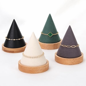 May include: Four cone-shaped jewelry display stands with wooden bases, featuring a black, cream, dark green, and gray velvet finish. Each stand holds a gold-toned bracelet, including a clover motif, a link chain, and two stone-set designs.