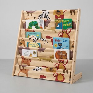 May include: A light wood bookshelf with five fabric tiers featuring animal illustrations. The shelves hold children's books including The Very Hungry Caterpillar, Goodnight Moon, The Pout-Pout Fish, Pete the Cat, and Brown Bear, Brown Bear, What Do You See?