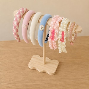 May include: A wooden display stand holding a collection of headbands and a beaded bracelet. The headbands include a pink gingham pattern, solid pink, white, blue with a floral emblem, and two ruffled fabric bands with the text Barbie.