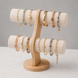 May include: A two-tiered light wood jewelry display stand with a beige velvet finish on the bars. Twelve various bracelets are arranged on the stand, including beaded, pearl, gold-tone chain, and hammered metal styles in assorted colors.