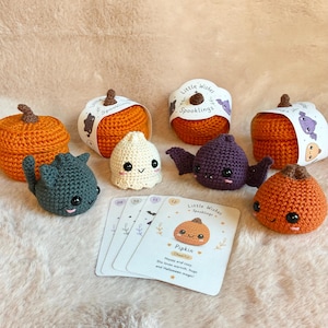 Op de afbeelding: Een verzameling gehaakte items waaronder oranje pompoenen met bruine steeltjes, een donkergrijze kat, een crèmekleurig spookje en een paarse vleermuis. Er liggen diverse kaarten, waarvan één met de tekst Little Wishes Spooklings erop.