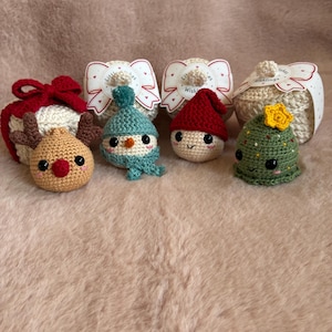 May include: Four small crocheted ornaments featuring a reindeer with brown antlers, a snowman in a blue hat, a gnome in a red hat, and a green tree with a yellow star. Each item is paired with a white gift box featuring a bow and the text Little Wishes.