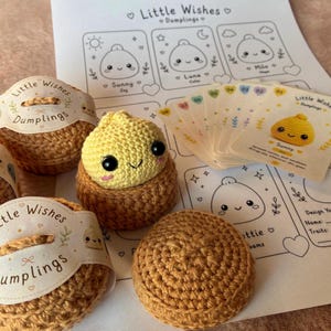 May include: A yellow crocheted dumpling toy with black eyes and pink cheeks, nestled in a brown crocheted base. Beside it are two brown crocheted containers with Little Wishes Dumplings paper labels and a set of illustrated character cards.