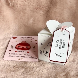 May include: A white gift box with a heart-shaped top closure and a tag reading A LITTLE SOMETHING FOR YOU. Beside it lies a square card featuring a cartoon dumpling character wearing a hat, with the text Little Wishes Dumplings Pippa Loves To Play And Making Friends.