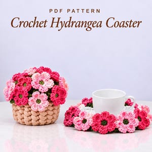 Peut inclure: Un modèle PDF intitulé Crochet Hydrangea Coaster montrant un panier crocheté beige rempli de fleurs d'hortensia roses et rouges, ainsi qu'un dessous de verre assorti supportant une tasse en céramique blanche.