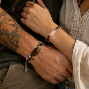 Puede incluir: Dos pulseras con bandas de cordón y detalles anudados. Una pulsera muestra una cuenta de piedra cilíndrica de color rosa claro suave, mientras que la otra presenta una cuenta de piedra ojo de tigre marrón pulida y curva con rayas oscuras.