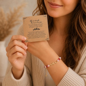 Puede incluir: Una pulsera de tono dorado con un dije central de ola y cuentas alternas rojas y rosa claro, acompañada de una tarjeta de papel kraft rectangular impresa con el texto: life is tough but so are you, junto a un icono de ola y un mensaje.