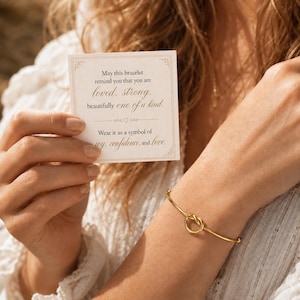 Puede incluir: Una pulsera rígida de metal dorado con un diseño de nudo central. Se sostiene una tarjeta blanca cuadrada cerca, impresa con el texto: May this bracelet remind you that you are loved, strong, beautifully one of a kind. Wear it as a symbol of joy, confidence, and love.