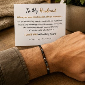 May include: A beaded bracelet featuring alternating dark brown, black, and metallic silver-toned cylindrical beads on a black cord. Behind the bracelet is a white card with black and gold text that reads To My Husband, When you wear this bracelet, always remember, You are the man of my dreams, my soul mate, and my other half. I feel so lucky for having you. I don't know anyone in this world who could love me with such passion and honesty. I can't imagine my life without you in it. I LOVE YOU with all my heart! followed by Morse code.