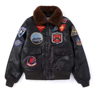 May include: A black leather bomber jacket featuring a brown faux fur collar, ribbed cuffs, and a hem. The jacket is adorned with various colourful embroidered patches, including a ship, a shield with the text U.S.S. WILLIAM H. STANDLEY, and aircraft.