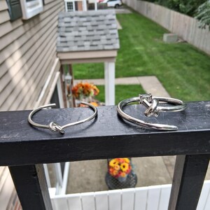 Może przedstawiać: Dwie srebrzyste bransoletki typu mankiet, każda z wyraźnym węzłem. Lewa bransoletka jest cieńsza z prostym węzłem, podczas gdy prawa jest grubsza i zawiera element w kształcie litery T zintegrowany ze strukturą węzła.