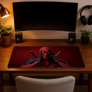 May include: A rectangular desk mat featuring a graphic illustration of the character Deadpool in a red and black suit with white eyes, positioned against a dark red background. The character has hands raised to the sides of the head.