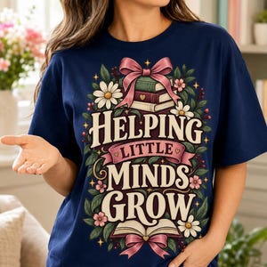 Könnte beinhalten: Ein marineblaues T-Shirt mit einem Grafikdruck und dem Text "HELPING LITTLE MINDS GROW". Das Design zeigt einen Bücherstapel, ein offenes Buch, rosa Schleifen, weiße Gänseblümchen und grüne Blätter in einem dekorativen Blumenrahmen.