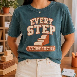 May include: A dark teal short-sleeve t-shirt featuring the text "EVERY STEP" in large cream letters above a graphic of a staircase. Below the stairs, a rectangular orange banner contains the text "LEARNING TOGETHER" in cream-coloured letters.