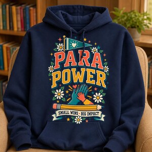 May include: A navy blue hooded sweatshirt featuring a graphic print with the text PARA POWER and SMALL WINS BIG IMPACT. The design includes a pennant, flowers, stars, a pencil, and two hands high-fiving in orange and teal colours.