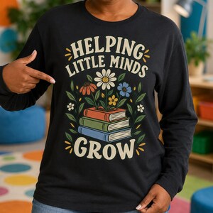 Könnte beinhalten: Ein schwarzes Langarmshirt mit einer Grafik von drei gestapelten Büchern, aus denen Blumen wachsen. Der Text "HELPING LITTLE MINDS GROW" ist in einer geschwungenen, cremefarbenen Schrift gedruckt. Das Design enthält grüne Blätter und bunte Blumen.