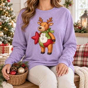 May include: A heathered light purple long-sleeve sweatshirt featuring a graphic of a brown stuffed reindeer toy with a green scarf, a red star, and a patchwork-style belly with two white buttons. The reindeer has stitched details.