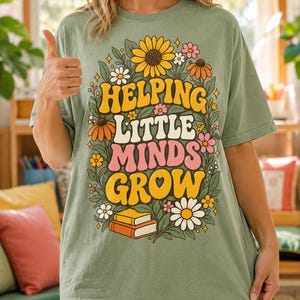 Könnte beinhalten: Ein salbeigrünes T-Shirt mit einem Grafikdruck aus bunten Blumen, Blättern und zwei gestapelten Büchern. Der Text "HELPING LITTLE MINDS GROW" ist mittig in einer fetten Retro-Schriftart mit gelben, weißen und rosafarbenen Buchstaben aufgedruckt.